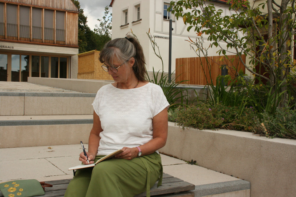 Kathrin Tröster in einem weißen lockeren T-Shirt und einer olivfarbenen Hose sitz auf einer Bank in der Niederwerrner 'Neuen Mitte'. Sie schreibt gerade etwas mit einem Stift in ein Buch.
